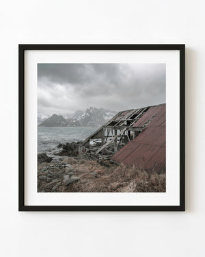 A decaying Lofoten boathouse with a broken red roof stands beside snowy Norwegian mountains and sea.