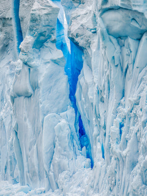 Folded white ice and soft shadows surround a narrow saturated blue fissure in Antarctica.
