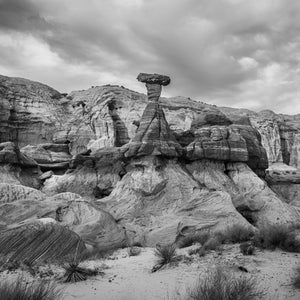 Narrow rock column rising above layered desert slopes under a cloudy sky.