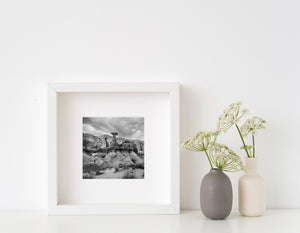 Layered cliffs and sparse plants surrounding a desert hoodoo, black and white.