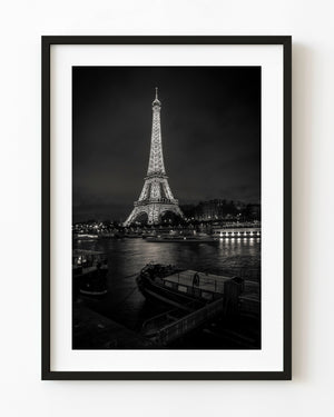 Illuminated Eiffel Tower above the Seine and shadowed river boats in black and white