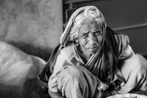 Elderly Indian woman seated with her knees drawn up in a black and white photograph.