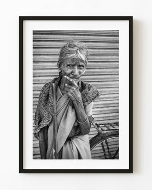 Elderly woman with a walking stick and raised hand in a black and white India street portrait.