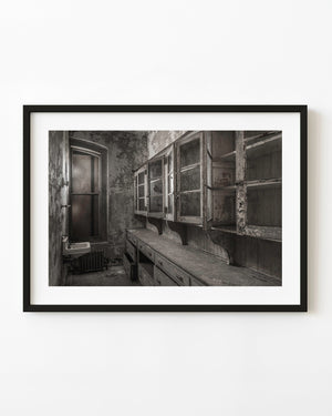 Open cupboards and a long counter inside Ellis Island Hospital’s abandoned stock room in black and white.