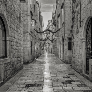 Cobbled street flanked by stone buildings with hanging lights in a black and white photo.