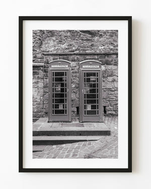 Two English telephone booths stand against a stone wall in black and white.