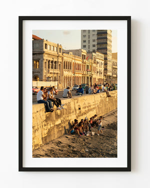 People gather along Havana’s seawall beneath sunlit waterfront buildings in Cuba.