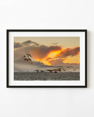 Orange and gold sunset light glows behind a snow-covered Antarctic mountain above dark water.
