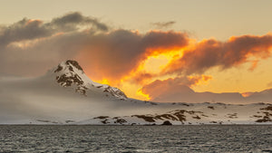 Snowy Antarctic peaks rise beneath grey clouds lit by a fiery sunset.