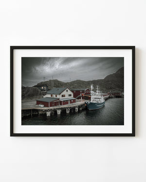 A green fishing vessel moored beside red waterfront buildings at Mortsund harbour in Lofoten, Norway.