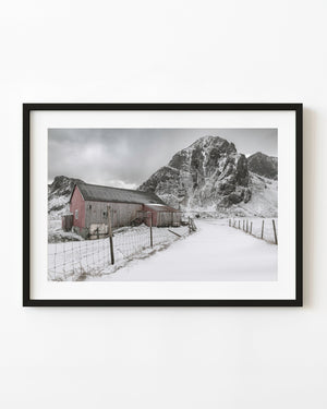 A weathered barn beside a snow-covered lane beneath a rugged mountain in Flakstad, Norway.