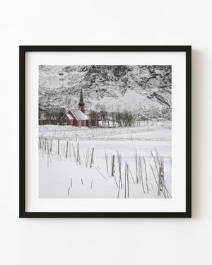 Red timber Flakstad Chapel stands in deep snow beneath a rugged Norwegian mountain.