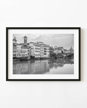 Florence buildings and Ponte Santa Trinita reflect in the Arno River, with a small boat, in black and white.