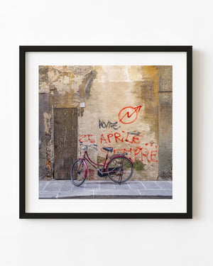A burgundy bicycle rests against a weathered Florence wall marked with red graffiti beside an old wooden door.