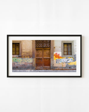 An old brown double door stands between barred windows on a graffiti-marked Florence wall in a colour photograph.