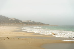 A foggy Australian shoreline with pale turquoise water, white foam and sandy dunes.