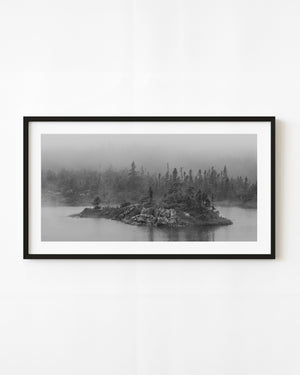 A rocky evergreen-covered island sits in a foggy Newfoundland lake in black and white.