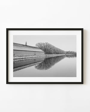 The Forbidden City outer wall and bare trees reflect in a calm Beijing moat in black and white.