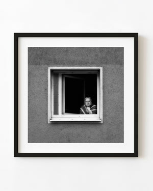 An older woman wearing glasses looks from an open Frankfurt window in a black and white portrait.