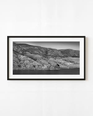 A long freight train follows the river beneath rocky Canadian hills in black and white