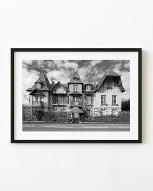 An ornate French mansion with steep tiled roofs and timber details beneath cloudy skies in black and white