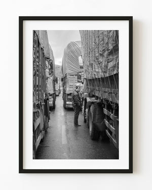 A man looks back between stopped freight trucks on a Ganzi highway in black and white