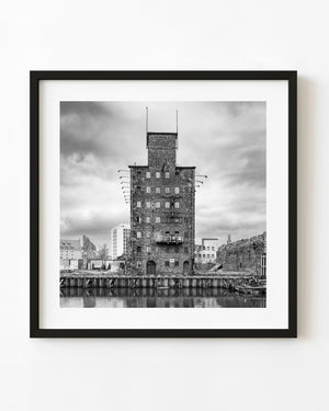 An abandoned brick riverfront tower stands beside calm water in Gdańsk, photographed in black and white