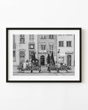 A coffee shop terrace with closed umbrellas and planters stands before historic Gdańsk façades in black and white