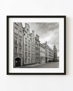 Gabled buildings line a cobblestone street in Gdańsk beneath a cloudy sky in black and white