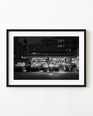 A brightly lit fruit and vegetable market stands beneath dark Gdańsk buildings in black and white