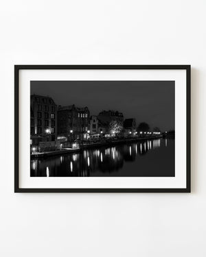 Riverfront lamps reflect in calm water beside Gdańsk buildings in a black and white night photograph