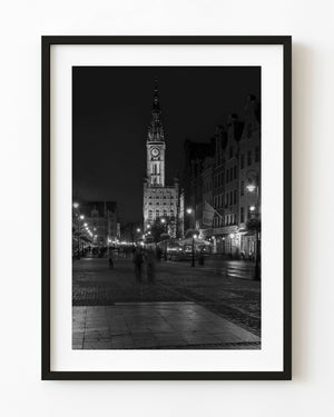 Gdańsk’s illuminated town hall rises above wet cobblestones and pedestrians in a black and white night photograph