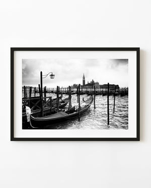 Gondolas rest beside wooden mooring posts on the Venice waterfront in black and white