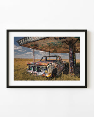 A graffiti-covered abandoned truck rests beneath a weathered Texaco canopy in Texas