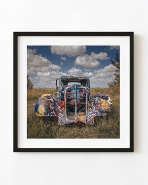 A graffiti-covered vintage truck stands in Texas grass beneath a cloud-filled blue sky