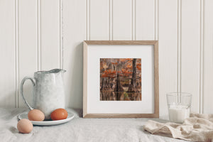 Framed artwork on a table with eggs and a pitcher against a white paneled wall.