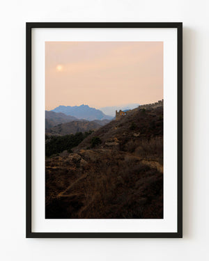 The Great Wall crosses dark hills beneath a hazy peach-coloured sunset sky