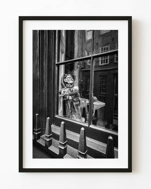 A grinning puppet appears behind a London window with reflected brick buildings in black and white