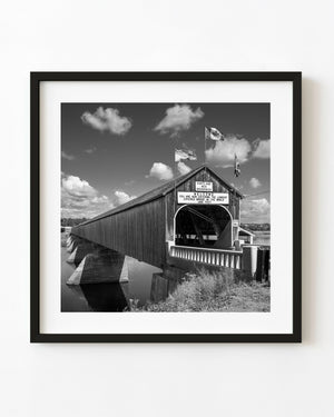 Hartland Covered Bridge spans a New Brunswick river beneath flags and clouds in black and white
