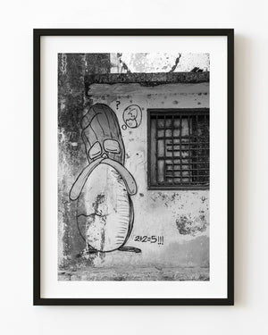 Cartoon graffiti beside a barred window on a weathered Havana wall, photographed in black and white.