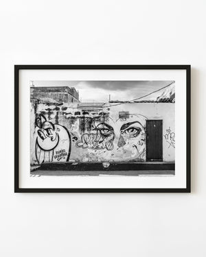 Large graffiti eyes and a cartoon figure cover a Havana wall beside a dark doorway in black and white.