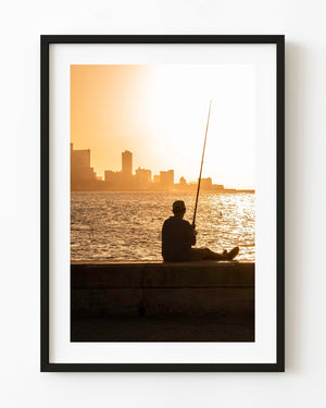 Fisherman silhouetted on Havana’s Malecon seawall beside golden water and the distant city skyline.