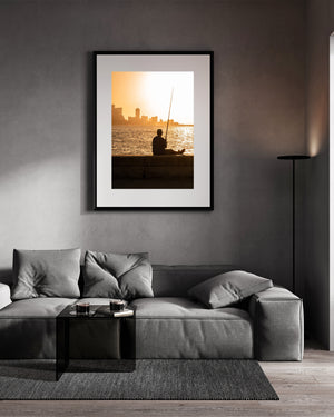 Colour photograph of a fisherman resting on the Malecon as sunset glows across the water.