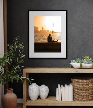A fisherman sits on Havana’s waterfront wall beneath an orange sky and distant buildings.
