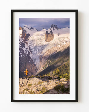 Hiker in an orange jacket photographs a glacier beneath rugged snowy Canadian mountain peaks.