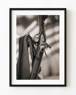 Sepia close-up of a historic musket’s metal lock and wooden stock with an American flag blurred behind.