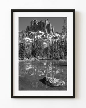 Howser Spire’s jagged peaks reflect in a small alpine pond beneath evergreen trees in black and white.