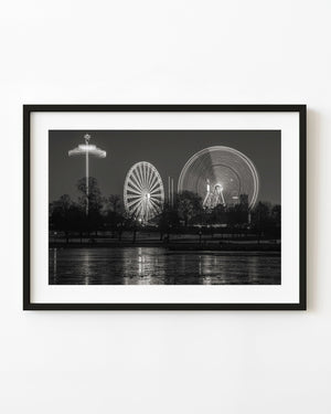 Illuminated fairground rides at Hyde Park Winter Wonderland, reflected across the lake in black and white.