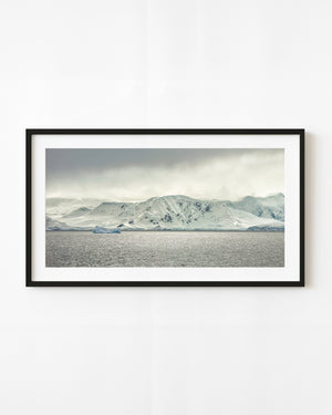 Ice-covered Antarctic mountains above a grey sea, with a small blue iceberg beneath heavy clouds.