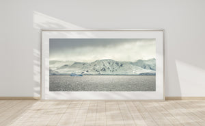 Light filters through clouds over snowy Antarctic ridges and a calm, textured sea.